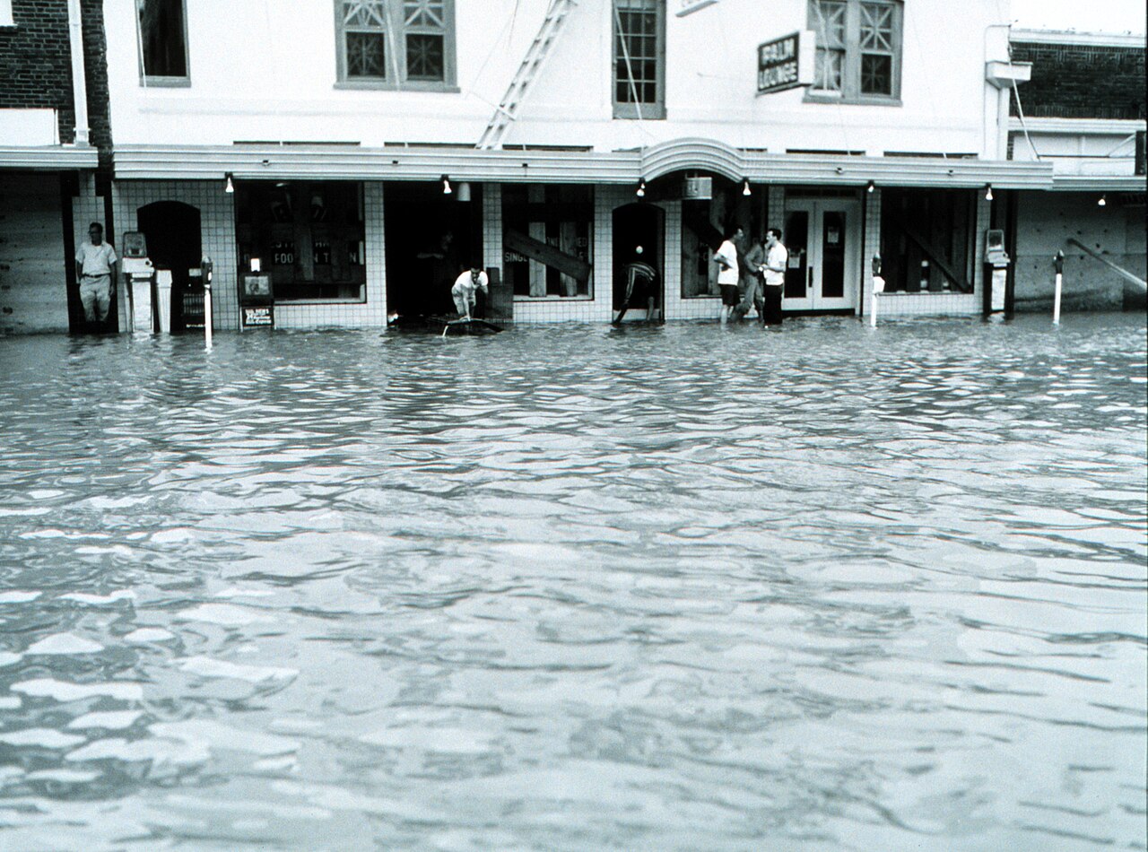 Black and white 1967 photograph of a flooded Rio Grande Valley street, with floodwater reaching the storefront awnings and several people standing in doorways